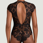 Black lace bodysuit shown from the back on a model, featuring a large open back, cap sleeves, and a matching black waist tie, set against a solid light gray background.
