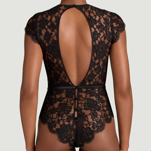 Black lace bodysuit shown from the back on a model, featuring a large open back, cap sleeves, and a matching black waist tie, set against a solid light gray background.