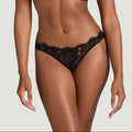 A model wearing a black lace lingerie set with a sheer floral lace bralette and matching lace thong, shown against a solid light gray background.