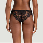 Rear view of a model wearing a black lace thong and matching black bra against a light gray studio background.