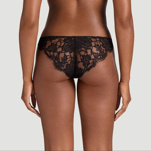 Rear view of a model wearing a black lace thong and matching black bra against a light gray studio background.