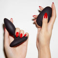 Black silicone butt plug held by two hands with red nail polish on a white background