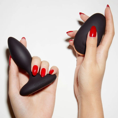 Black silicone butt plug held by two hands with red nail polish on a white background